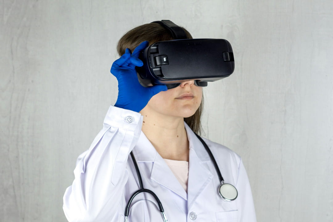 Young woman shown from chest up, she is dressed in a white lab coat and blue gloves. She has a stethoscope around her neck and a virtual reality headset over her eyes.